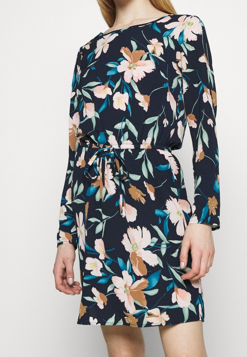 Femme portant une robe florale bleu marine à manches longues et une ceinture nouée, se tenant devant un fond clair et uni.