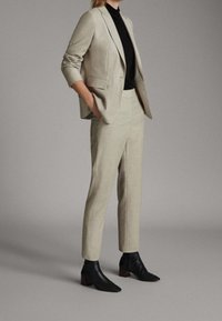 Blazer beige ajusté avec des épaules structurées, associé à un pantalon assorti. Un col roulé noir en dessous et des bottines noires complètent la tenue.
