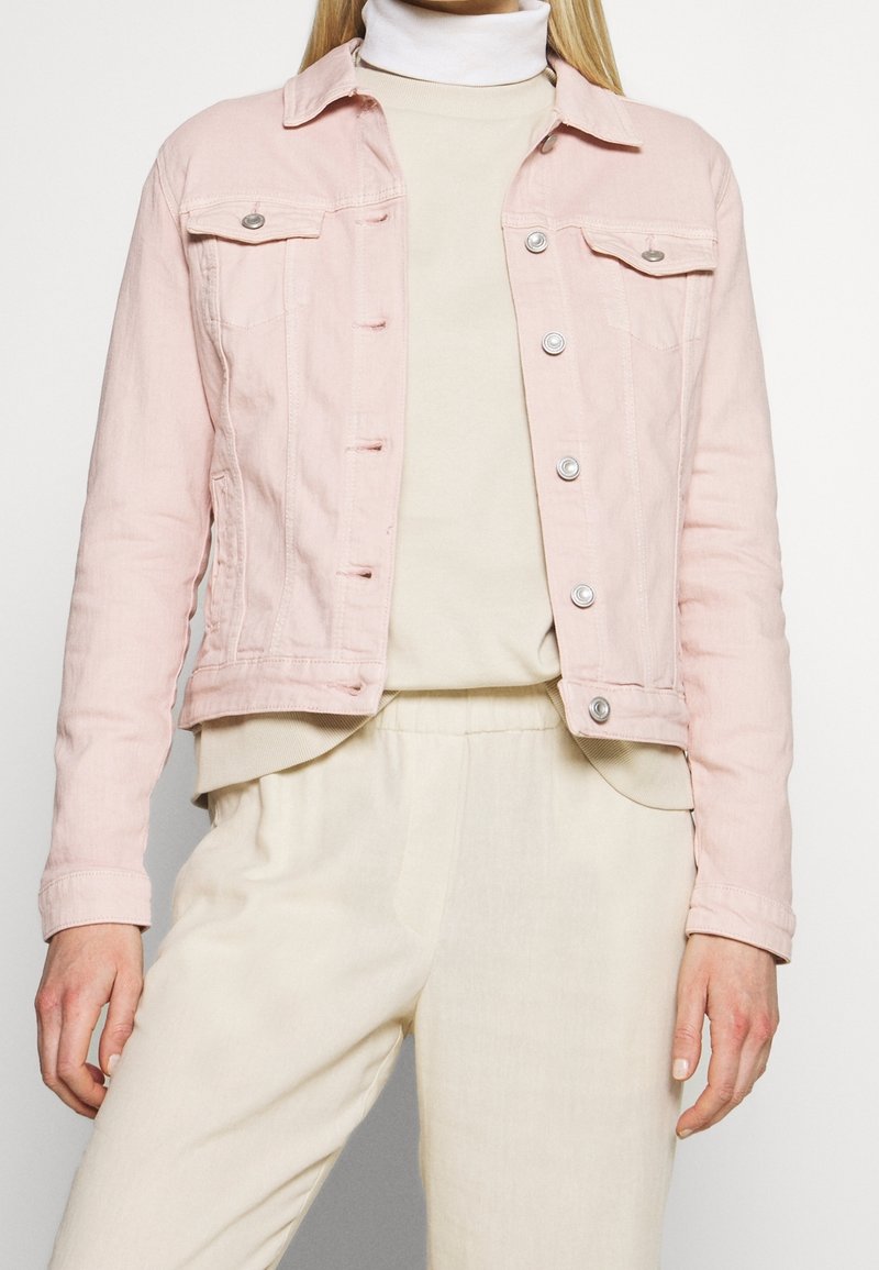 Femme portant une veste en jean rose clair sur un pull à col roulé crème et un pantalon crème assorti, debout sur un fond uni.