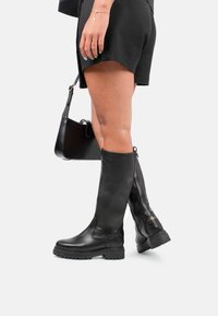 Botas de cuero negras hasta la rodilla con suela gruesa, cremallera lateral y textura suave. Combinadas con pantalones cortos negros y un bolso pequeño.