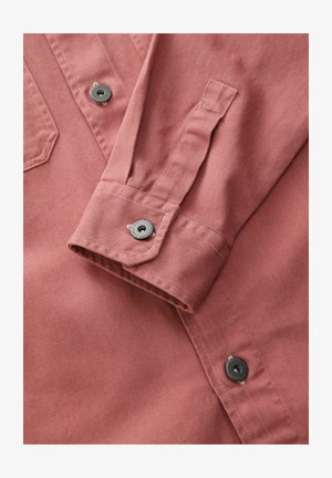 Piros pamut ing textúrált felülettel, gombos mandzsettával és fekete gombokkal; tartalmaz egy zsebet és ferde varrás részleteket.