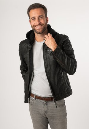 Giacca di pelle nera con cappuccio removibile, chiusura con zip e tasche laterali, abbinata a una t-shirt grigia chiara e jeans grigi.