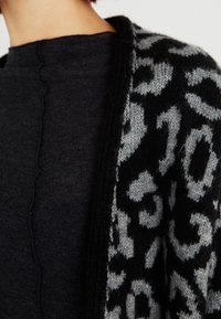 Cardigan de punto en negro y gris, con un patrón de leopardo. La tela es suave, con un frente abierto y un acabado texturizado.