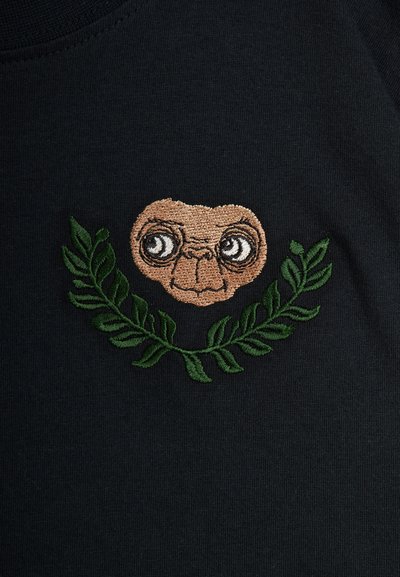 Melna auduma T-kreklis ar izšūtu rakstu, kas attēlo varoņa seju bēšā krāsā, ko ieskauj zaļi lapu akcenti.