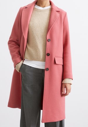 Frau trägt einen kniehohen rosa Wollmantel über einem beigen Pullover und grau gestreiften Hosen, mit einer Hand in der Tasche und der anderen locker entspannend.