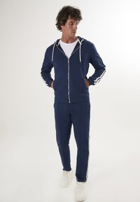 Chándal navy hecho de una tela suave; cuenta con una sudadera con capucha y cremallera con cordones blancos y franjas laterales, acompañado de pantalones ajustados y zapatillas blancas.