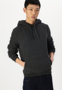 Sudadera con capucha de felpa en color carbón con acabado texturizado, capucha con cordón, bolsillo tipo canguro y puños y dobladillo acanalados. Llevada con jeans oscuros.
