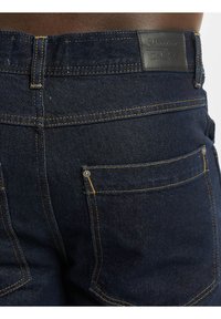 Temno modri denim kavbojki z gladko teksturo, rumenim šivom in usnjenim označevalnim delom na pasu z blagovno znamko.