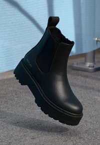 Black leather platform ankle boot with elastic sides and pull tab, branded "Replay," positioned mid-air above asphalt surface.