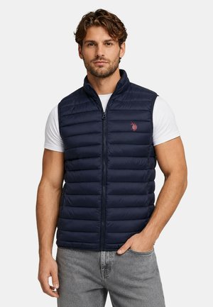 Man with short curly hair wears navy blue quilted vest over white t-shirt and gray jeans, standing with one hand in pocket against plain background.