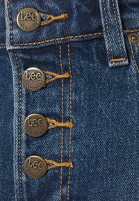 Tessuto denim con cinque bottoni metallici incisi con "Lee Est. 1889." Presenta cuciture gialle e un colore blu classico. Vista ravvicinata.