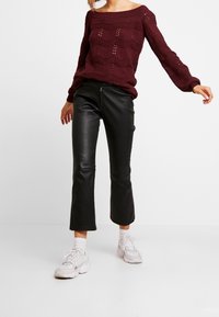 Bordowy sweter dzianinowy z wzorem w zygzak i otwartym splotem, zestawiony z czarnymi, skórzanymi spodniami w wersji cropped oraz białymi sneakerami.