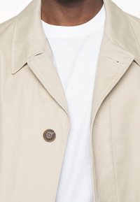 Nahaufnahme einer Person, die einen beigen Trenchcoat über einem einfachen weißen T-Shirt mit Rundhalsausschnitt trägt.