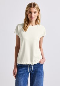 Chemise en crème claire, texturée, à manches courtes, avec un col rond et un ourlet réglable à cordon de serrage, associée à un jean bleu à jambes larges.