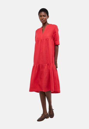 Femme portant une robe midi rouge ample avec un col en V et des manches aux coudes, associée à des chaussures Mary Jane en tresse marron foncé.