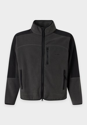 Homme aux cheveux tressés portant une veste noire et grise, un t-shirt blanc, un pantalon noir et des chaussures noires avec des semelles beiges, debout sur un fond blanc.