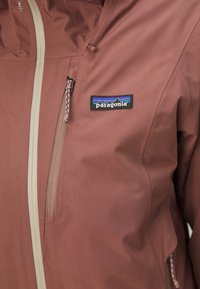Vandafvisende brun Patagonia jakke med en beige lynlås og lomme, samt et logo patch på brystet og en glat tekstur.