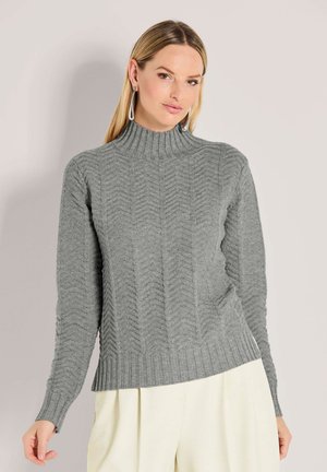 Vrouw in een grijsgenaakte gebreide trui met een hoge geribbelde kraag en crèmekleurige wijde broek tegen een effen lichte achtergrond.