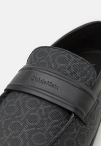Mocassins noirs à enfiler avec un dessus en tissu texturé arborant un motif de logo discret. Une bande noire lisse avec la marque "Calvin Klein" accentue le design.