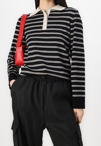 Kvinna som bär en svart och vit randig långärmad polotröja, svarta cargobyxor med elastisk midja och bär en röd axelväska.