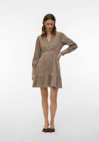 Femme portant une robe taupe à manches longues, avec des boutons sur le devant et un ourlet à volants, debout avec une main sur la hanche.