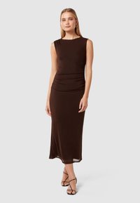 Robe marron sans manches en tissu élastique, dotée d'une taille froncée et d'une silhouette longue et fluide. Associée à des sandales noires à lanières.