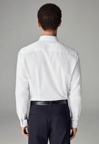 Cămașă albă cu mânecă lungă, cu o croială ajustată, având un guler clasic și manșete cu butoni, asortată cu pantaloni negri și o curea neagră.