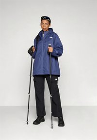 Person iført en blå North Face-jakke og svarte bukser, som holder svarte fjellstaver, står mot en ensfarget lys grå bakgrunn.