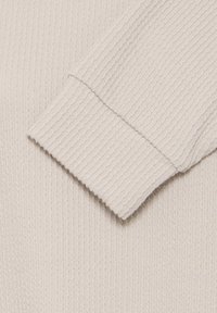 Gros plan sur la manche beige d'un pull tricoté montrant la texture côtelée et le détail du poignet replié.