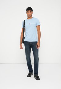 Šviesiai mėlynas medvilninis marškinėliai su "TOMMY HILFIGER EST. 1985" logotipu, derinami su tamsiai mėlynomis džinsais ir juodomis lengvomis bateliais, laikant juodą maišą.