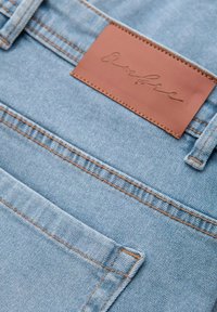 Ljust blå denimjeans har en sydd ficka, kontrasterande orange tråd och en brun läderetikett med handskriven stiltext.