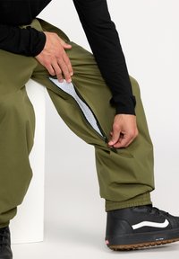 Olivegrüne, lässige Hose mit Netzfutter, die mit seitlichen Reißverschlusslüftungen und elastischen Bündchen ausgestattet ist, kombiniert mit schwarzen Schuhen.