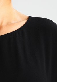 Sort bluse med en bred halsudskæring, blød tekstur og en glat overflade. Stoffet samles let ved skulderen for en afslappet pasform.