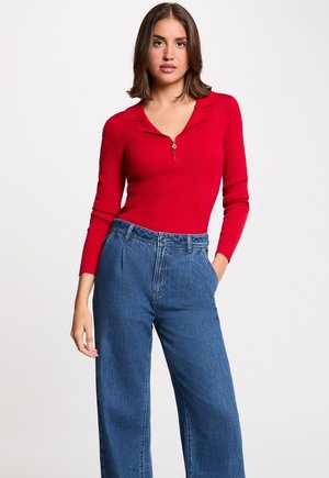 Kvinde iført en rød langærmet ribstrikket top med lynlås foran og blå høj talje vidde jeans, poserer med den ene hånd i lommen.