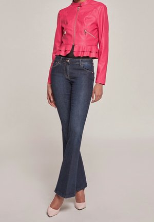 Femme portant une veste en cuir rose avec un ourlet à volants, un jean évasé bleu foncé et des talons pointus rose clair, debout devant un fond uni.