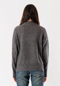 Persona con capelli lunghi castani che indossa un maglione grigio a maglia e jeans blu, in piedi rivolto di spalle davanti a uno sfondo bianco uniforme.