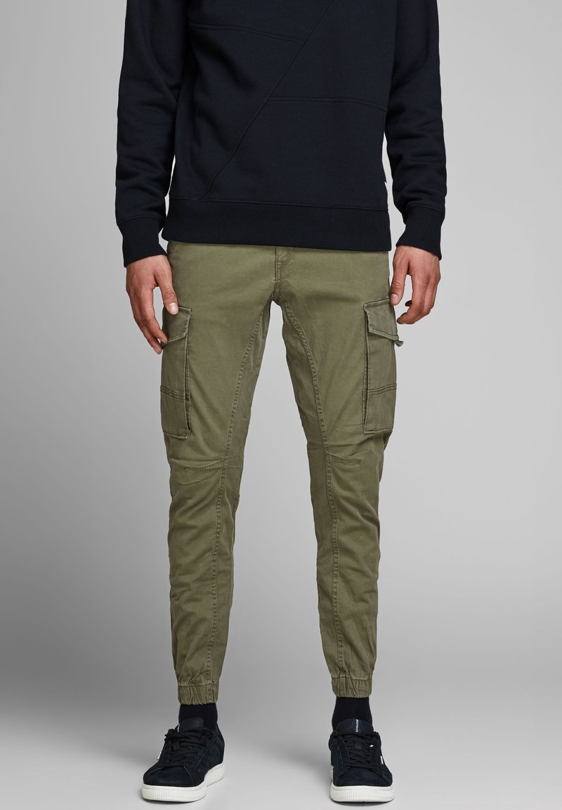 Homme portant un pantalon cargo jogger vert olive avec des poches latérales, un sweat-shirt noir et des baskets noires sur fond gris.