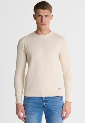 Cream-colored, textured knit pullover with a round neckline and ribbed cuffs, paired with blue jeans. Subtle logo detail at the hem.