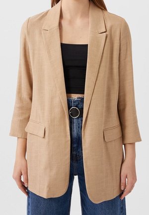 Woman wearing a beige blazer with 3/4 sleeves over a black crop top and blue high-waisted jeans with a black belt.