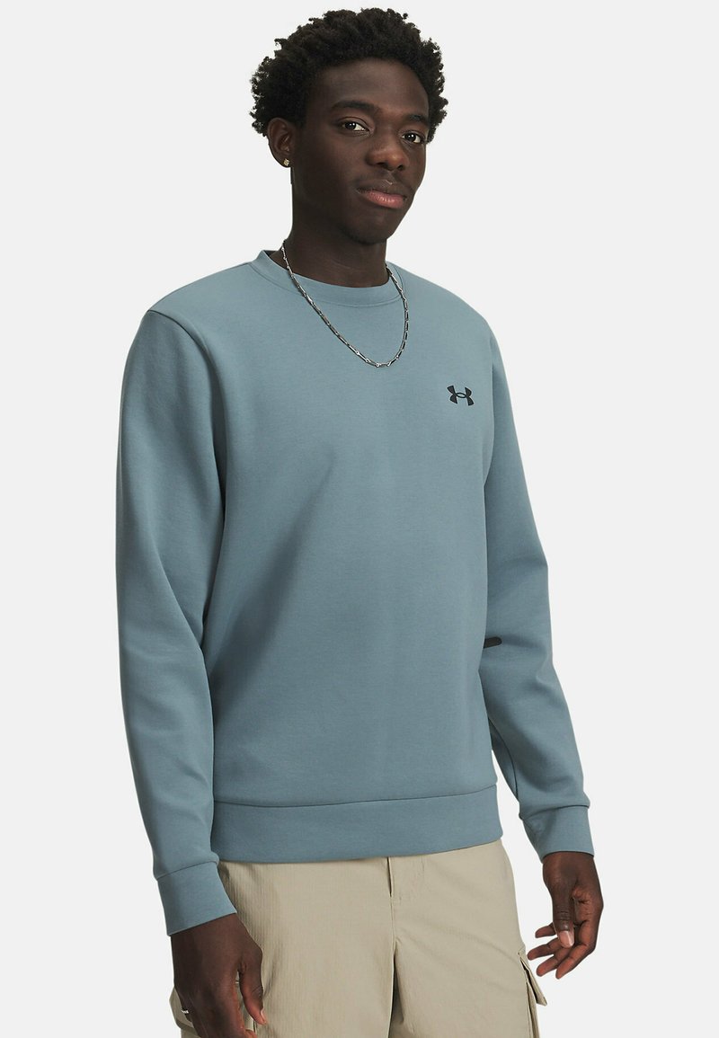 Young man stands confidently in a blue sweatshirt and beige shorts, with a silver chain necklace and a subtle logo on his chest.