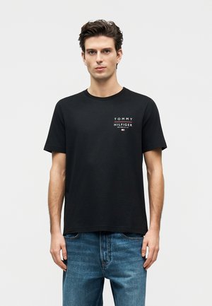 Mężczyzna w czarnej koszulce Tommy Hilfiger z logo na piersi, w połączeniu z niebieskimi dżinsami, stoi na jednolitym jasnym tle.