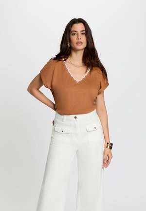 Donna con lunghi capelli scuri indossa una maglietta marrone a maniche corte con bordi bianchi e pantaloni bianchi a vita alta e gamba larga, in piedi con una mano sui fianchi.