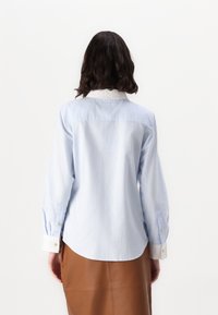 Donna con capelli scuri e mossi indossa una camicia azzurro chiaro con bottoni, con polsini e colletto bianchi, abbinata a una gonna di pelle marrone, in posizione di spalle.