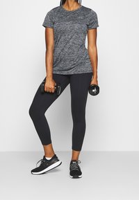 Szary melanżowy sportowy top z krótkim rękawem, czarne dopasowane legginsy, trzyma hantle oraz nosi czarne sportowe buty z białymi podeszwami.