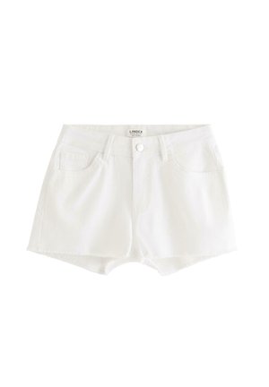 Teenagepige står iført en brun one-shoulder top med flæser, hvide denimshorts og sølvfarvede flade sko på en ensfarvet hvid baggrund.