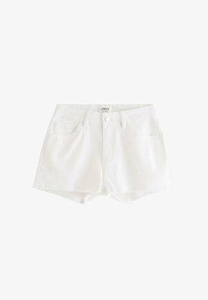 Teenagepige står iført en brun one-shoulder top med flæser, hvide denimshorts og sølvfarvede flade sko på en ensfarvet hvid baggrund.