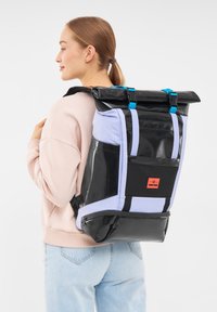 Hátizsák, amely fekete és világoslila anyagok kombinációjával készült, feltekerhető dizájnnal, műanyag részletekkel és egy kiemelkedő címkével az elején.