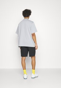 Muž s kudrnatými vlasy, oblečený v volné světle šedé košili s krátkým rukávem, černých šortkách, jasně žlutých ponožkách a bílých teniskách, stojí zády.