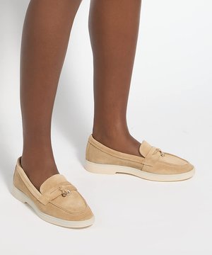 Jambes portant des mocassins en daim beige avec des semelles en caoutchouc clair et un petit détail de chaîne métallique sur fond blanc.