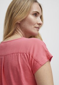Tričko s krátkým rukávem z jemné růžové látky s hladkou texturou, s mírným leskem a kulatým výstřihem, odhalující mírný záhyb na zadní straně.